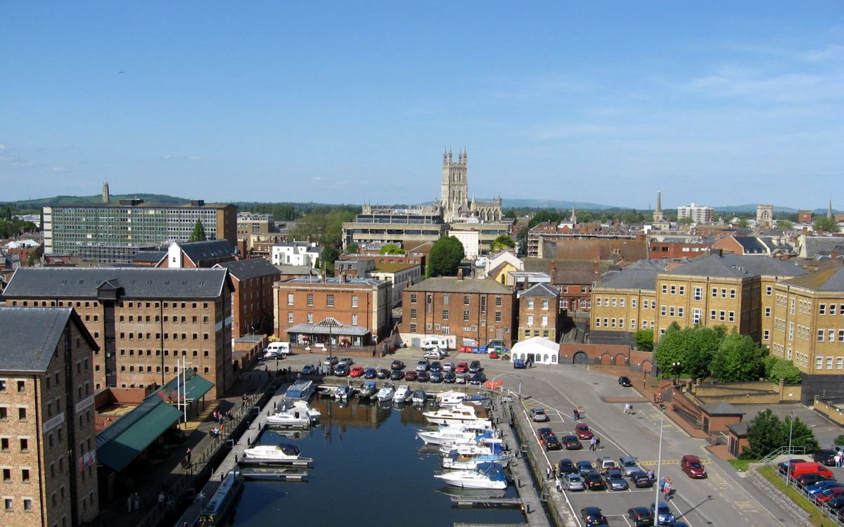 Gloucester Docks marina and brick warehouses seen from above, with Gloucester Cathedral tower on the skyline under blue sky