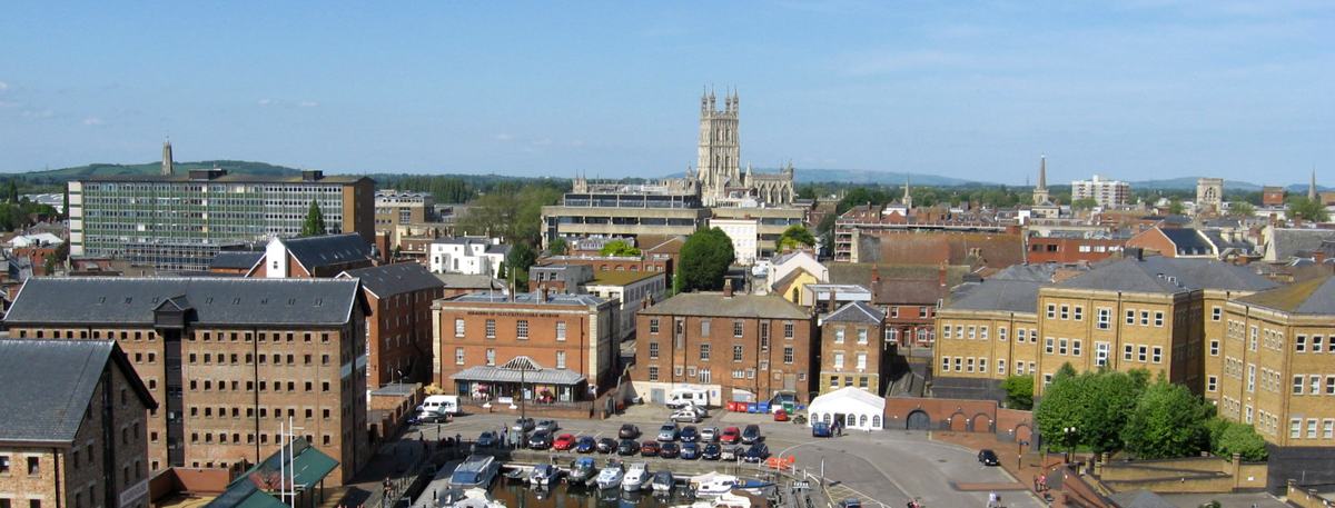 Gloucester Docks marina and brick warehouses seen from above, with Gloucester Cathedral tower on the skyline under blue sky