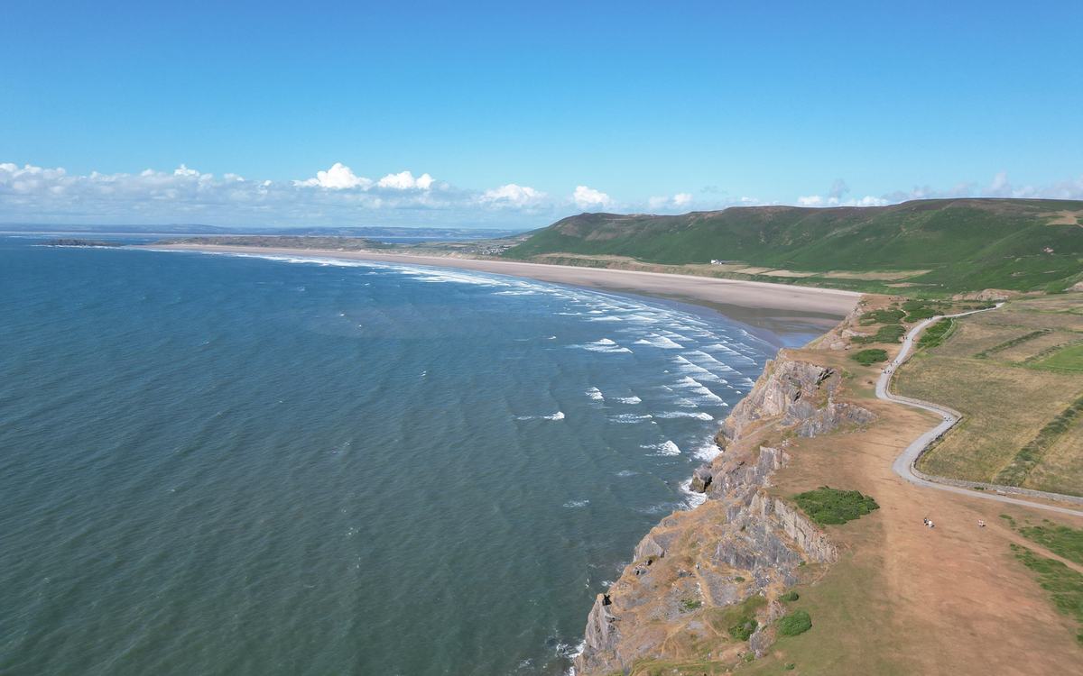 Drone view along the clifftops at Rhossili on the Gower Peninsula, with a long sandy bay, surf and green hills under blue sky