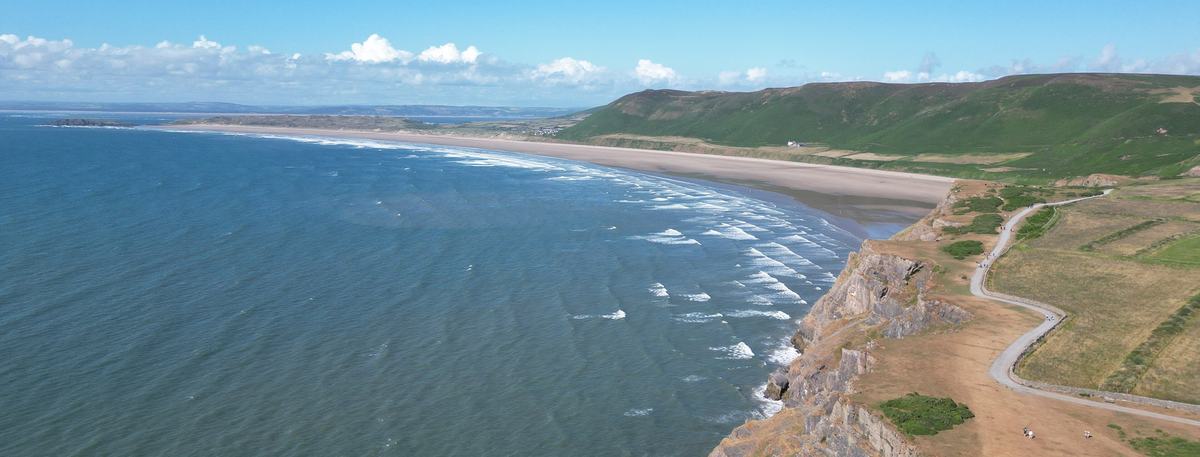 Drone view along the clifftops at Rhossili on the Gower Peninsula, with a long sandy bay, surf and green hills under blue sky