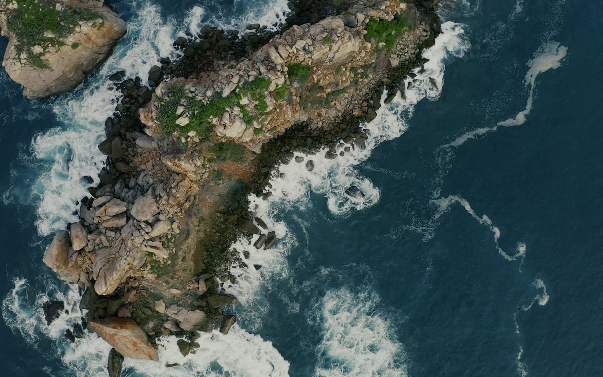 Top-down aerial view of a rocky islet with green patches, ringed by white surf in deep blue sea