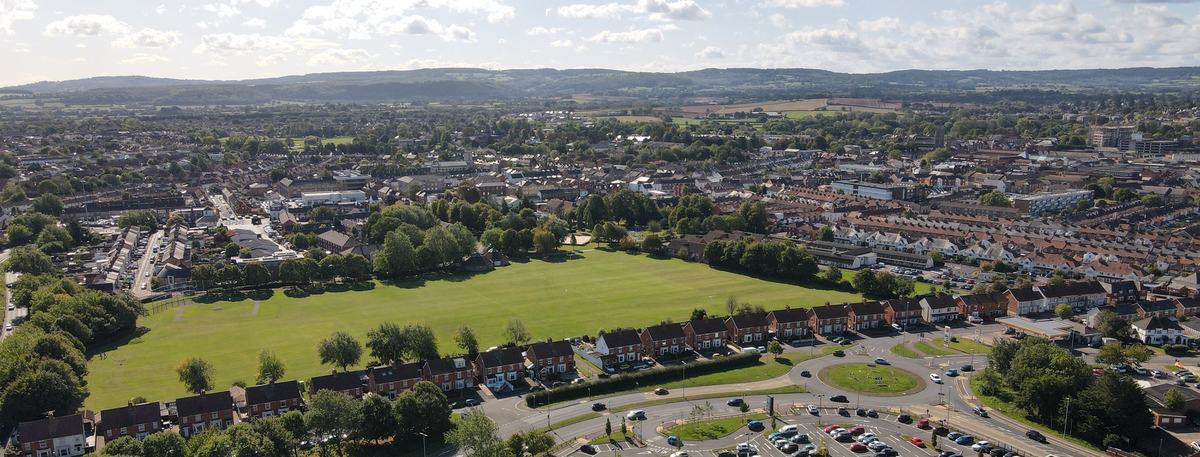 Drone view over Victoria Park in Taunton, with surrounding streets and wooded hills on the horizon under a blue sky