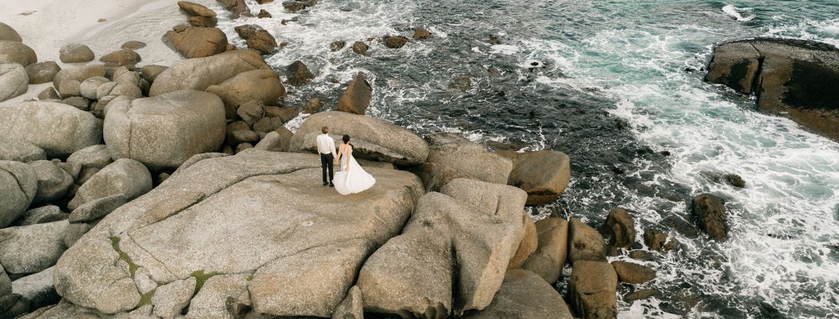 Drone view of a bride and groom standing on large granite boulders above a turquoise sea